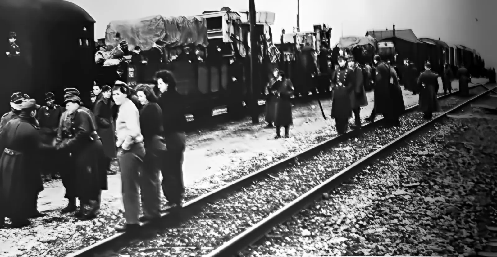 Menschen in Mänteln und Uniformen an den Gleisen neben einem langen Güterzug mit Ladung unter Planen