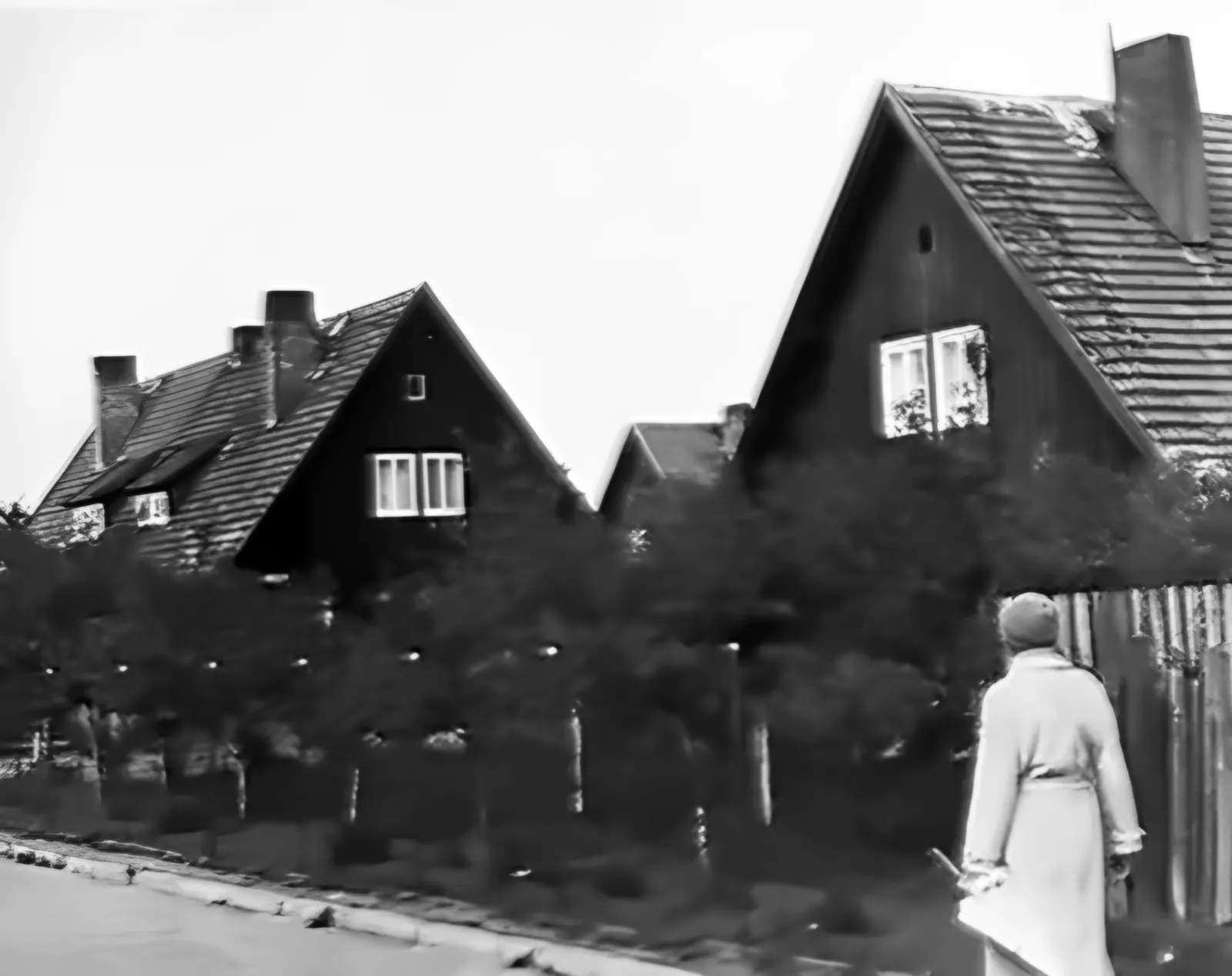 Holzhäuser mit Satteldächern hinter einer Hecke, auf dem Gehweg eine Frau im hellen Mantel